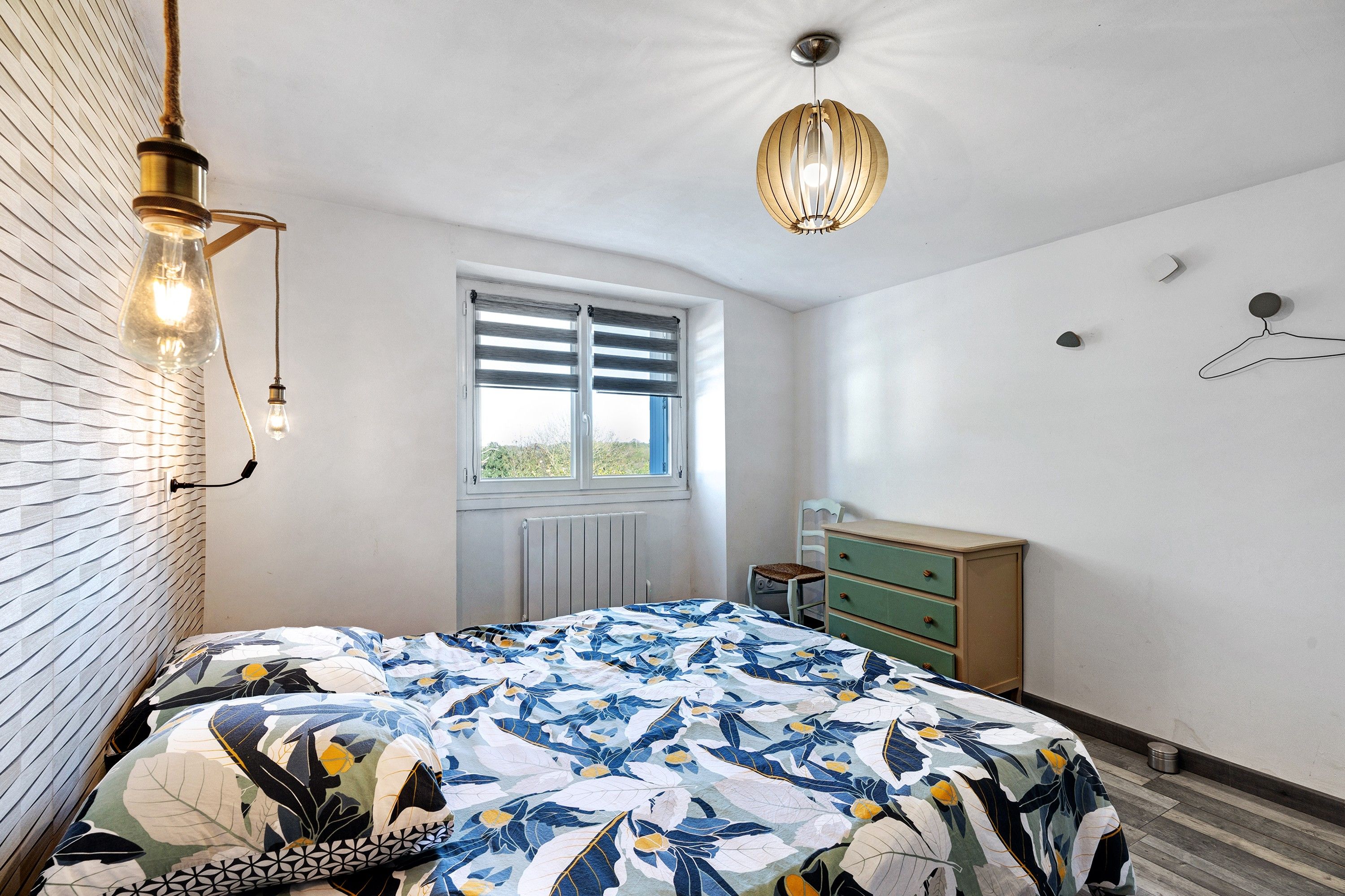 Ein Schlafzimmer mit Bett, Kommode, Fenster und Pendelleuchte.