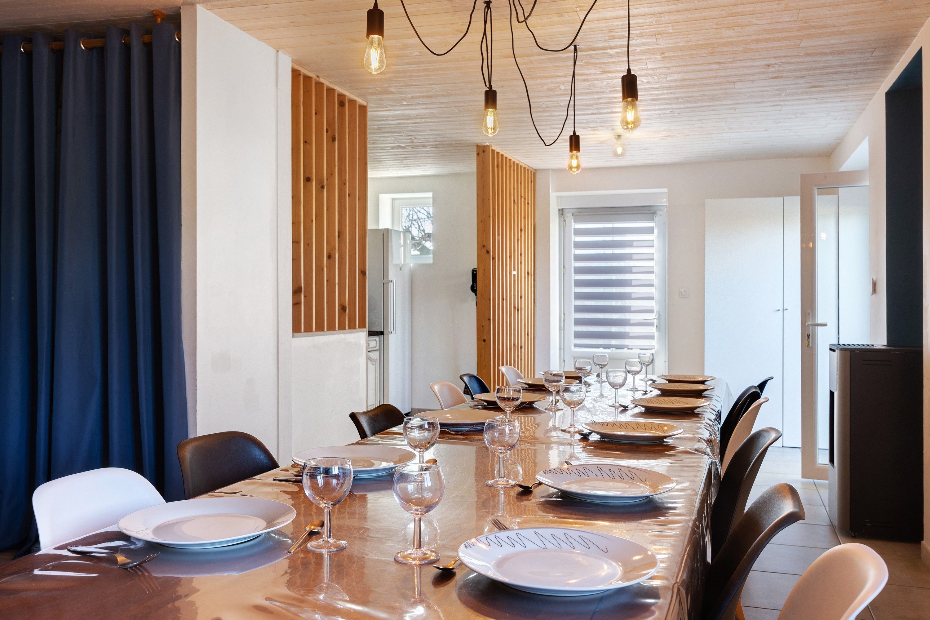 A dining area with a long table set for a meal, wooden paneling on the walls, and a ceiling with exposed beams and hanging lights.