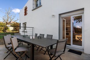 Un patio exterior con una mesa y sillas, una puerta corrediza de vidrio que da a una cocina, y un edificio blanco con una ventana y una pequeña estructura de ladrillo.