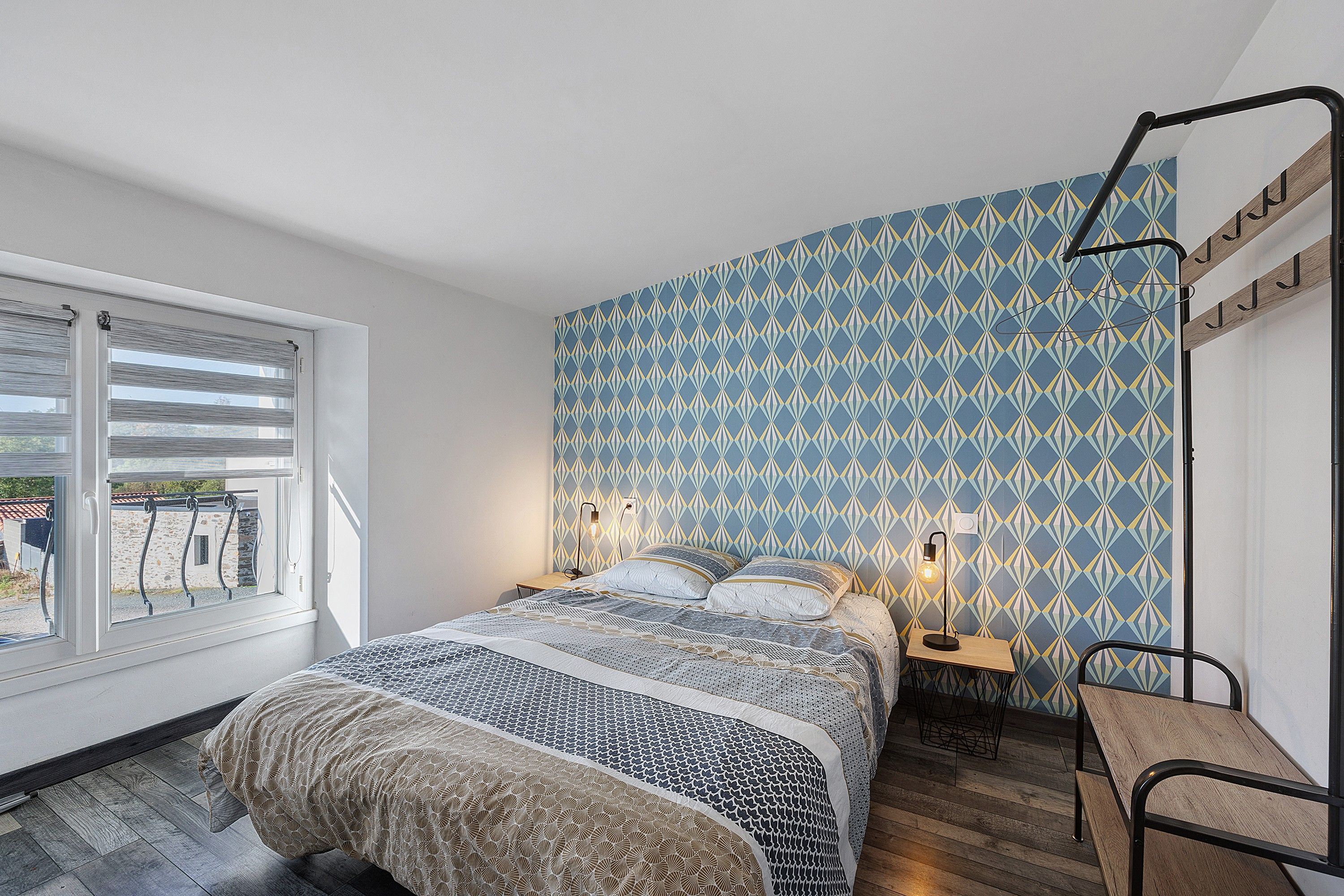 Ein Schlafzimmer mit gestreifter Wand, einem Bett mit gemusterten Betttextilien, einem Schreibtisch mit Stuhl und einem Fenster mit Jalousien.