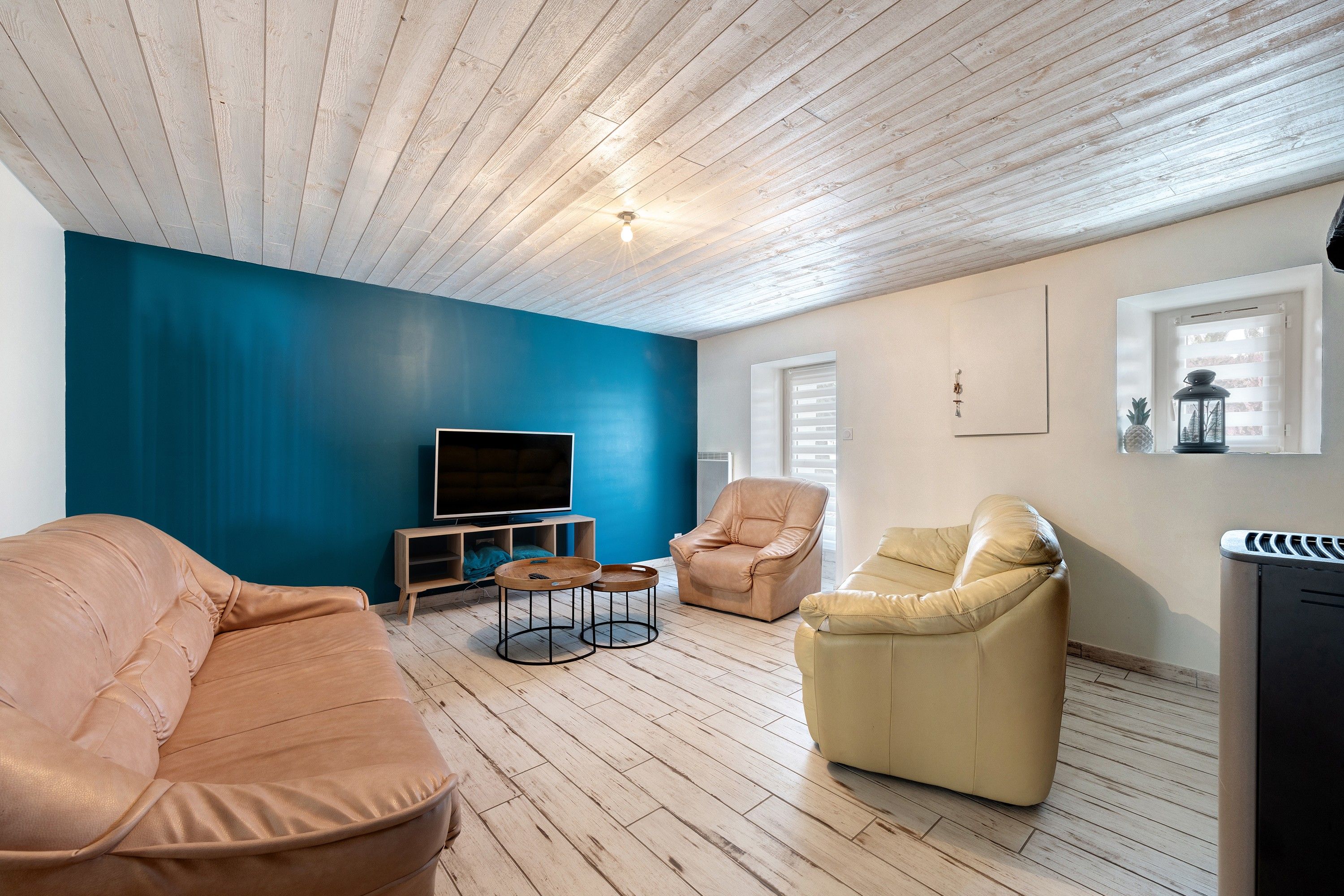 Una sala de estar con una pared azul, piso de madera, un televisor de pantalla plana, dos sofás de cuero y un ventanal con persianas.