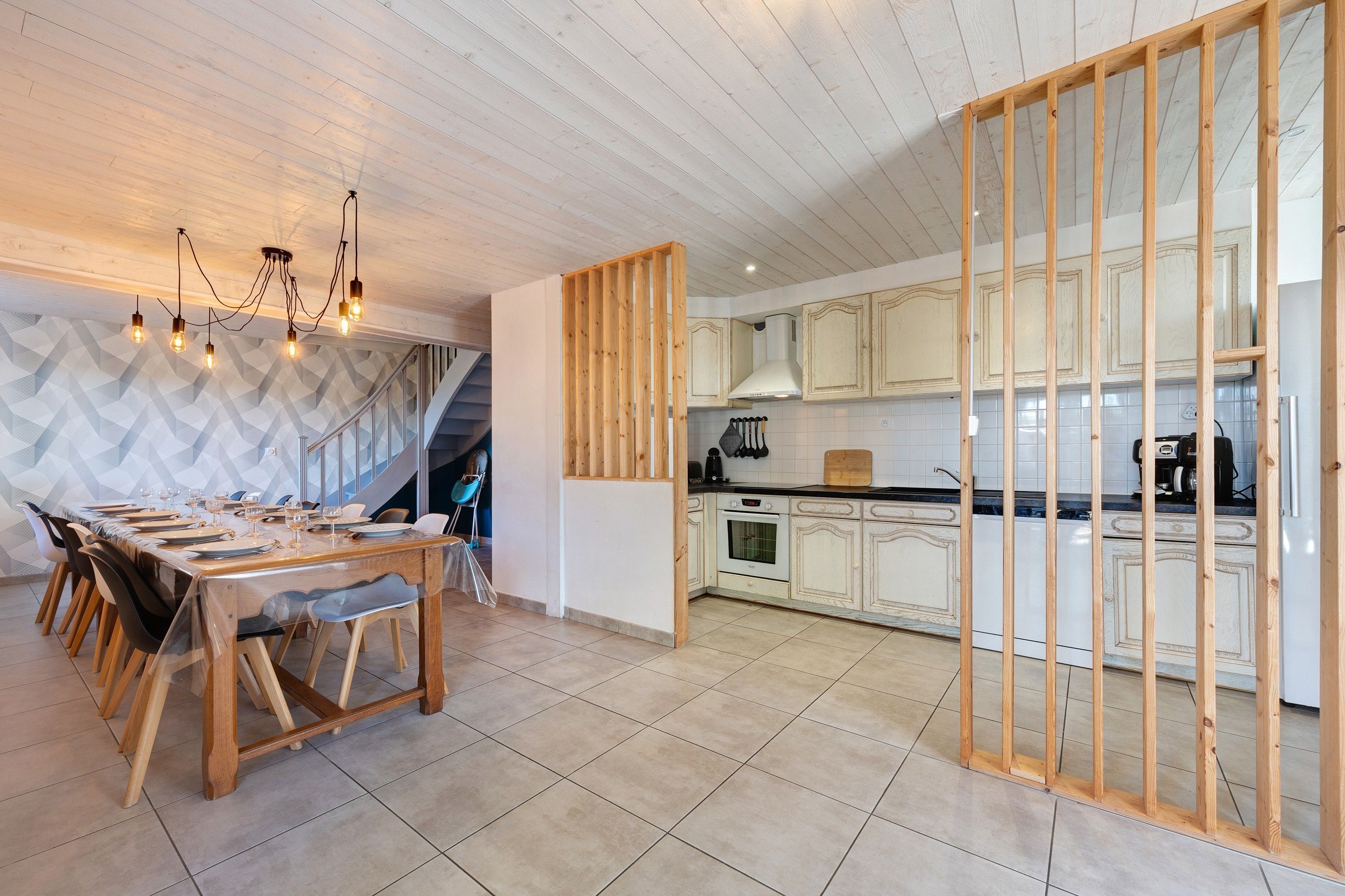 A modern kitchen with wooden cabinets, a dining area with a table set for four, and a staircase leading to an upper level.