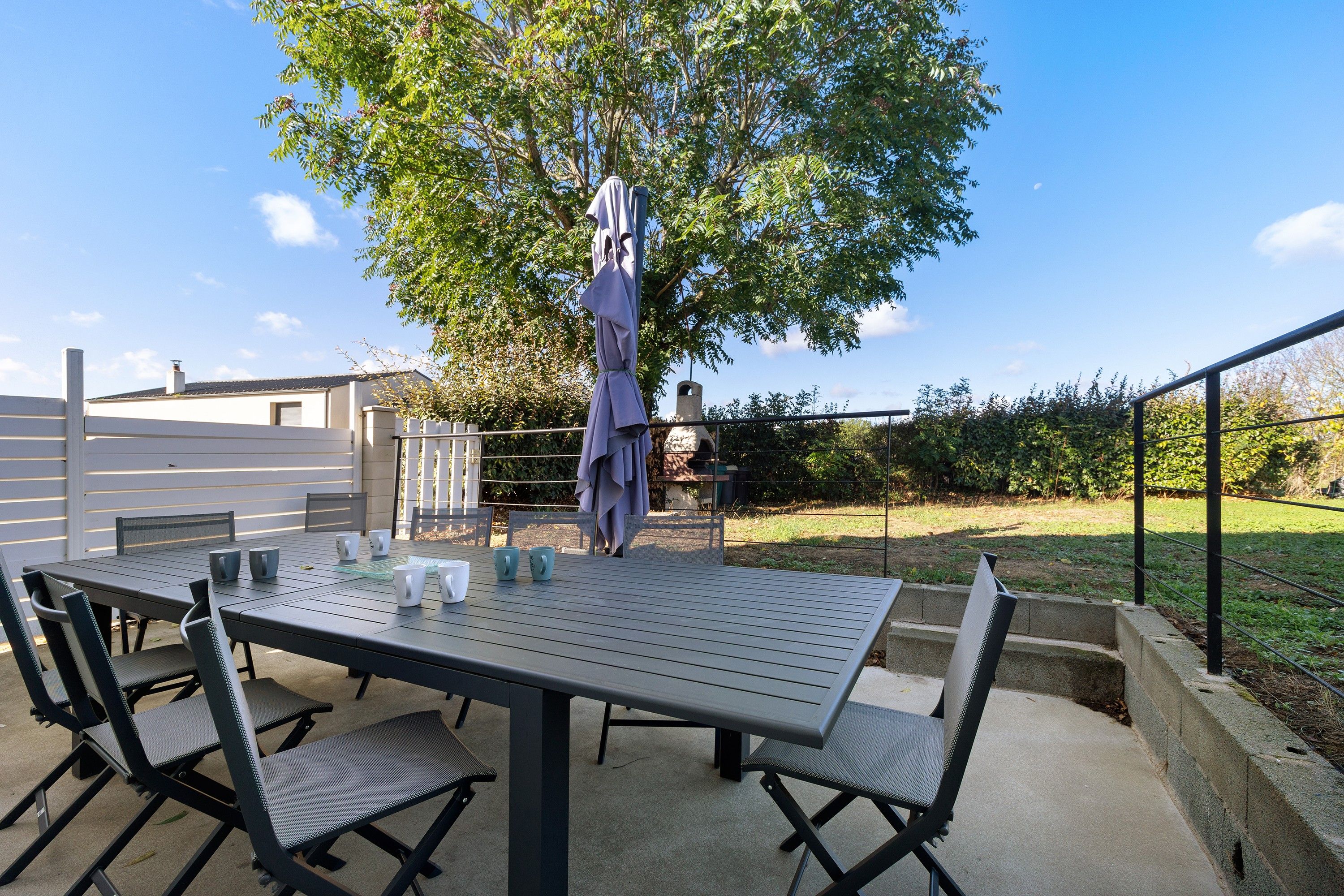 Eine Außenpatio mit Tisch und Stühlen, umgeben von einer Hecke und einem Baum.
