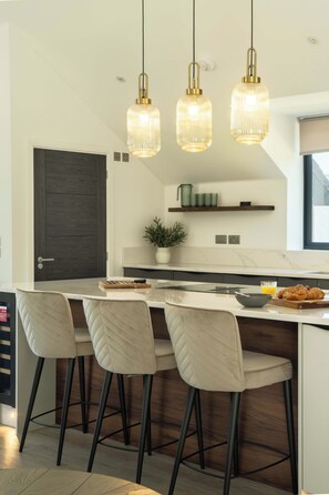 A modern kitchen with a central island, bar stools, pendant lighting, and a dark door. - Luttrell House - 3 bed eco house (Somerset)