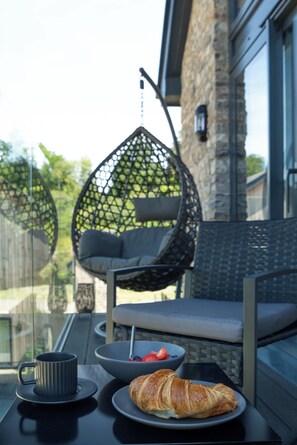 A balcony with a wicker chair, a table with a bowl of fruit and a croissant, and a mug. - Luttrell House - 3 bed eco house (Somerset)
