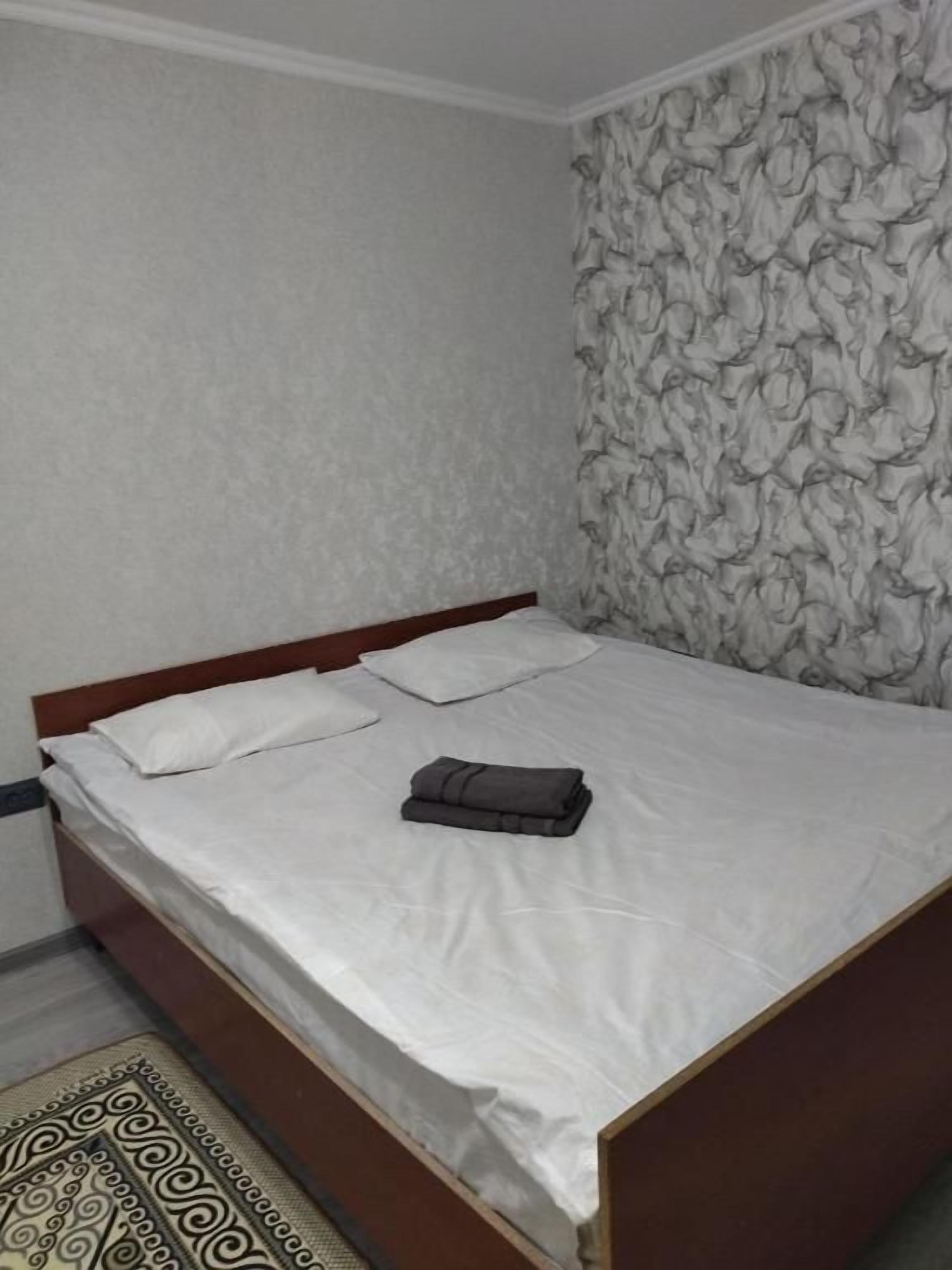 A single bed with a wooden frame and white bedding, a patterned wallpaper on the wall, and a patterned rug on the floor.