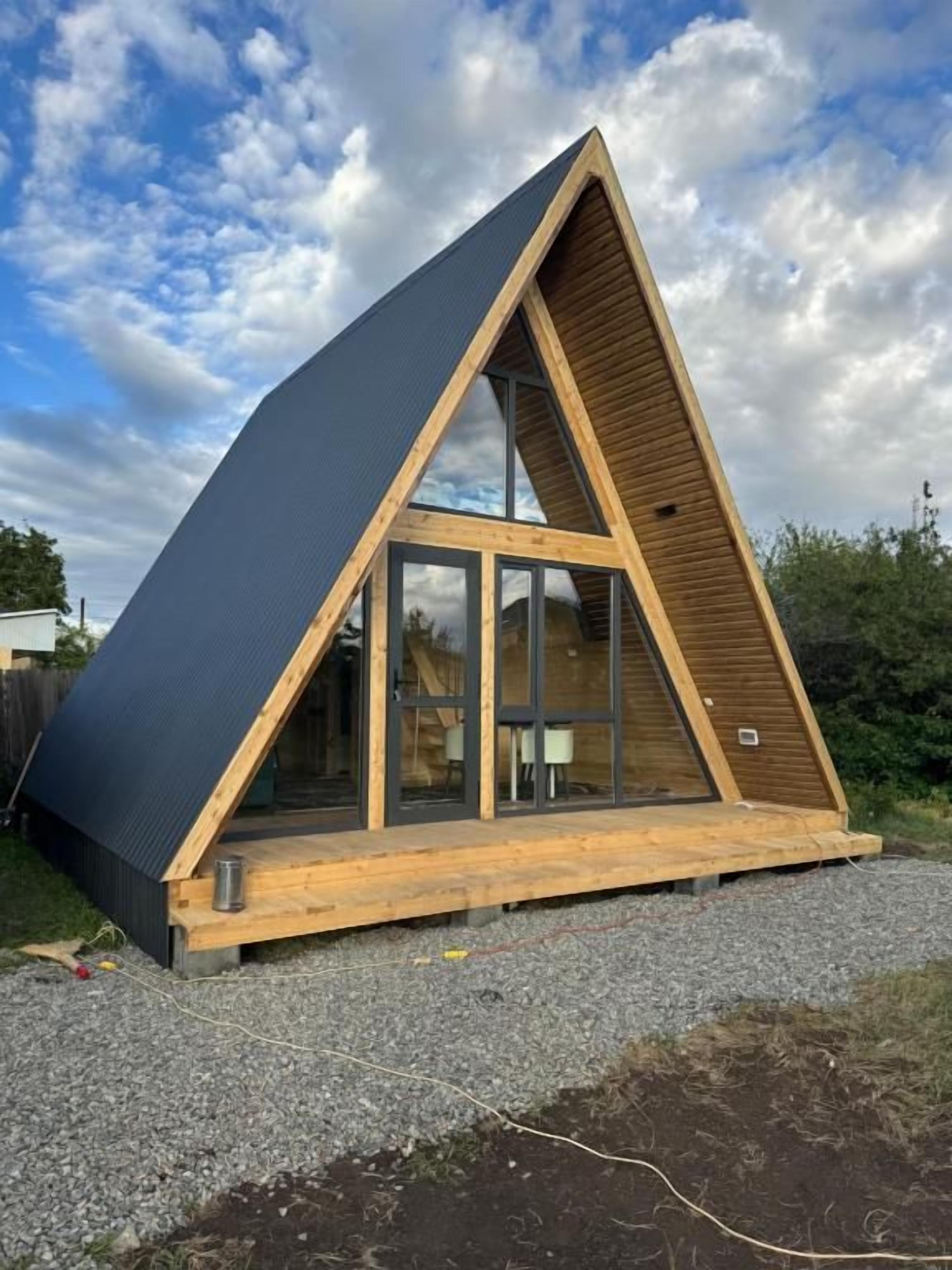 A triangular wooden structure with a glass facade, a flat roof, and a gravel ground.