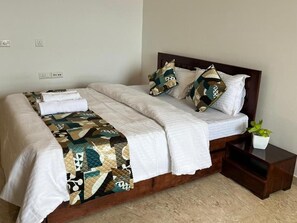 A neatly made bed with a quilt and pillows, a bedside table with a plant, and a power outlet on the wall. - Luna Spaces Riva (mumbai)
