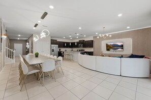 A modern kitchen with dark cabinets, a dining area with a marble table, and a living area with a white sofa and a mounted TV. - 3029 Magical Themed Home Dino Slide Pool Spa (Davenport)