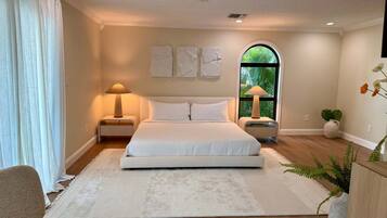 A bedroom with a large bed, two bedside tables with lamps, a window with a view of greenery, and a white rug.