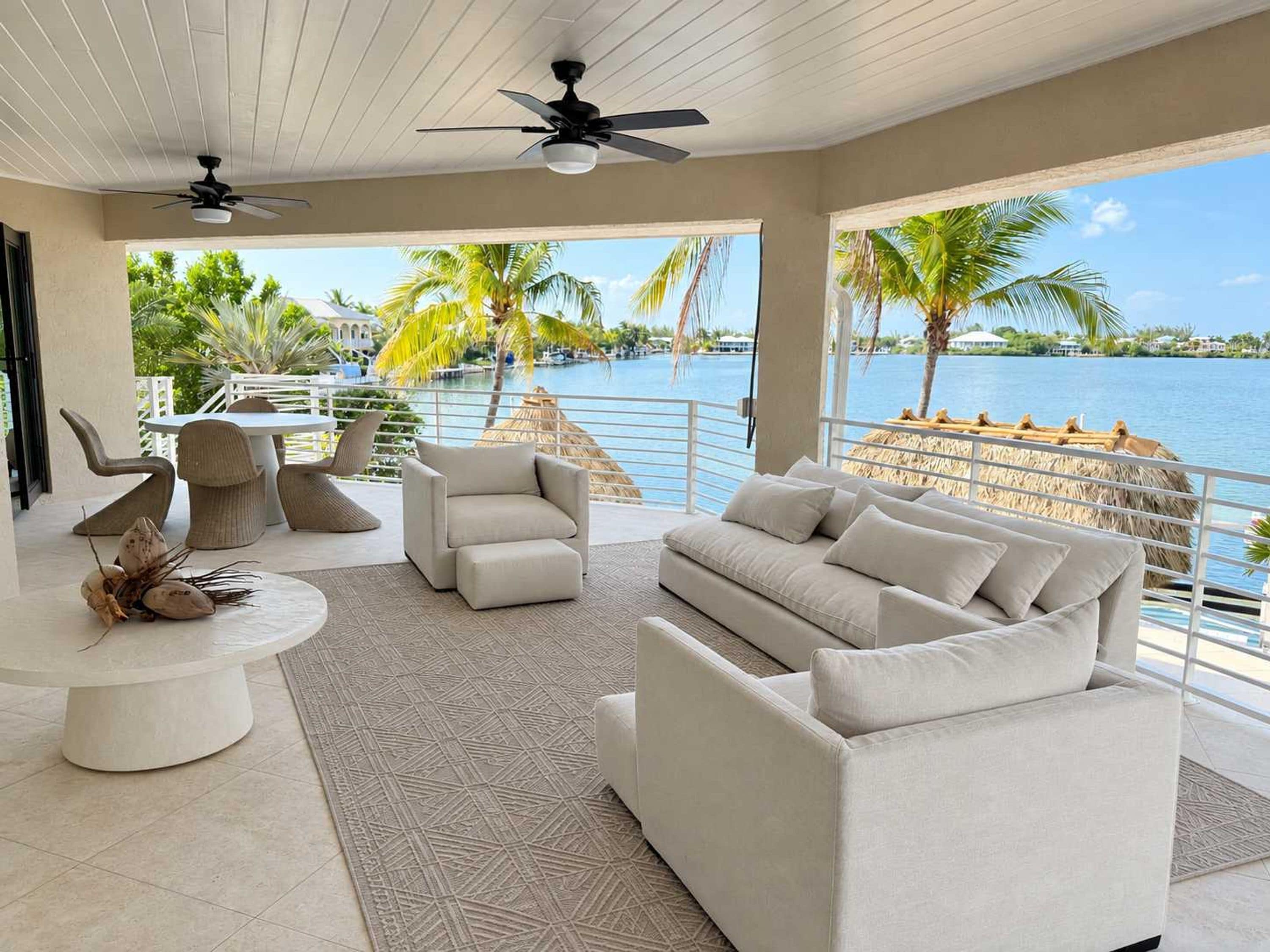 A modern living room with a view of a body of water, featuring a sofa, chairs, a dining table, and ceiling fans.