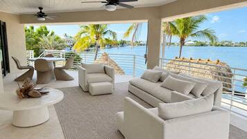 A modern living room with a view of a body of water, featuring a sofa, chairs, a dining table, and ceiling fans.