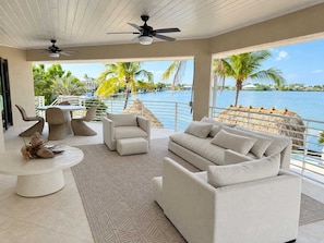 A modern living room with a view of a body of water, featuring a sofa, chairs, a dining table, and ceiling fans. - Kokomo Keys - Waterfront Heated Pool (Summerland Key)