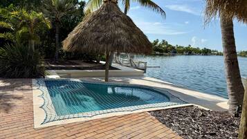 A swimming pool with a thatched roof structure, surrounded by palm trees and a body of water.