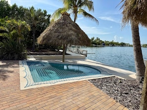 A swimming pool with a thatched roof structure, surrounded by palm trees and a body of water. - Kokomo Keys - Waterfront Heated Pool (Summerland Key)