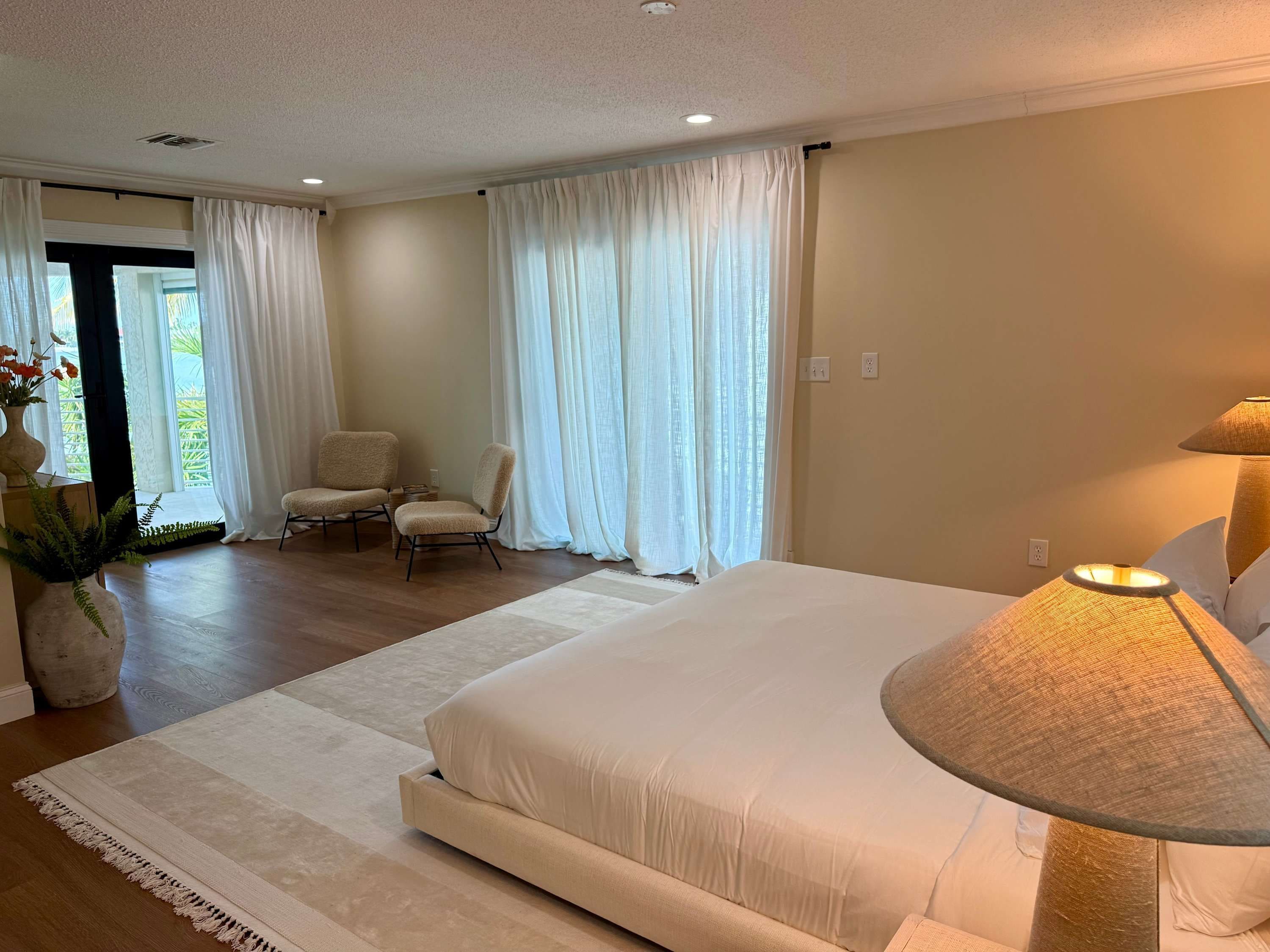 A bedroom with a large bed, two chairs, a lamp, and a view of greenery outside.