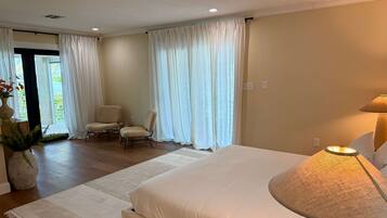 A bedroom with a large bed, two chairs, a lamp, and a view of greenery outside.