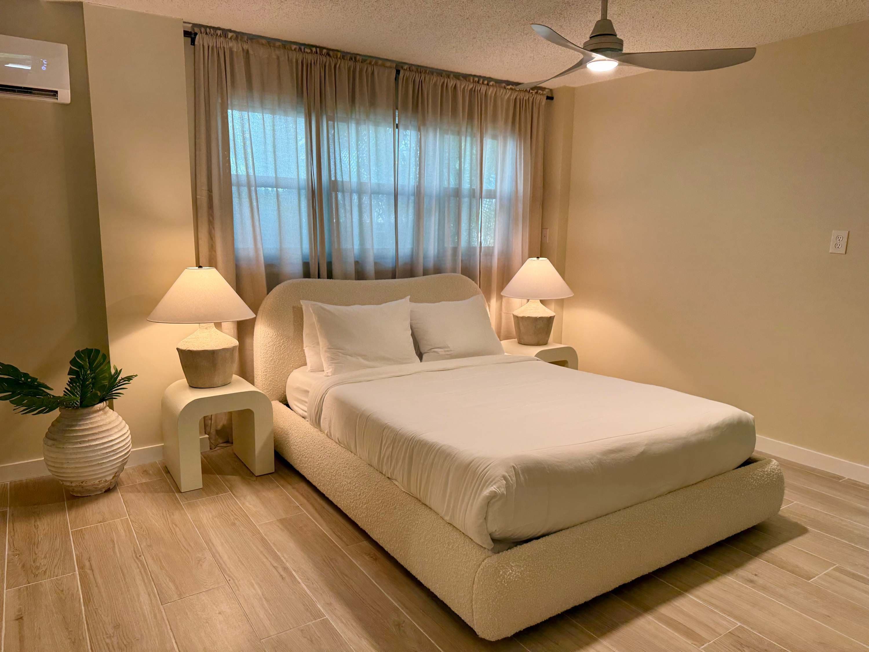 Une chambre à coucher avec un grand lit, deux lampes de chevet, un ventilateur de plafond et une fenêtre avec des rideaux.