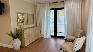 A hotel room with a wooden floor, a seating area with two chairs, a large window with curtains, and a wall-mounted TV.