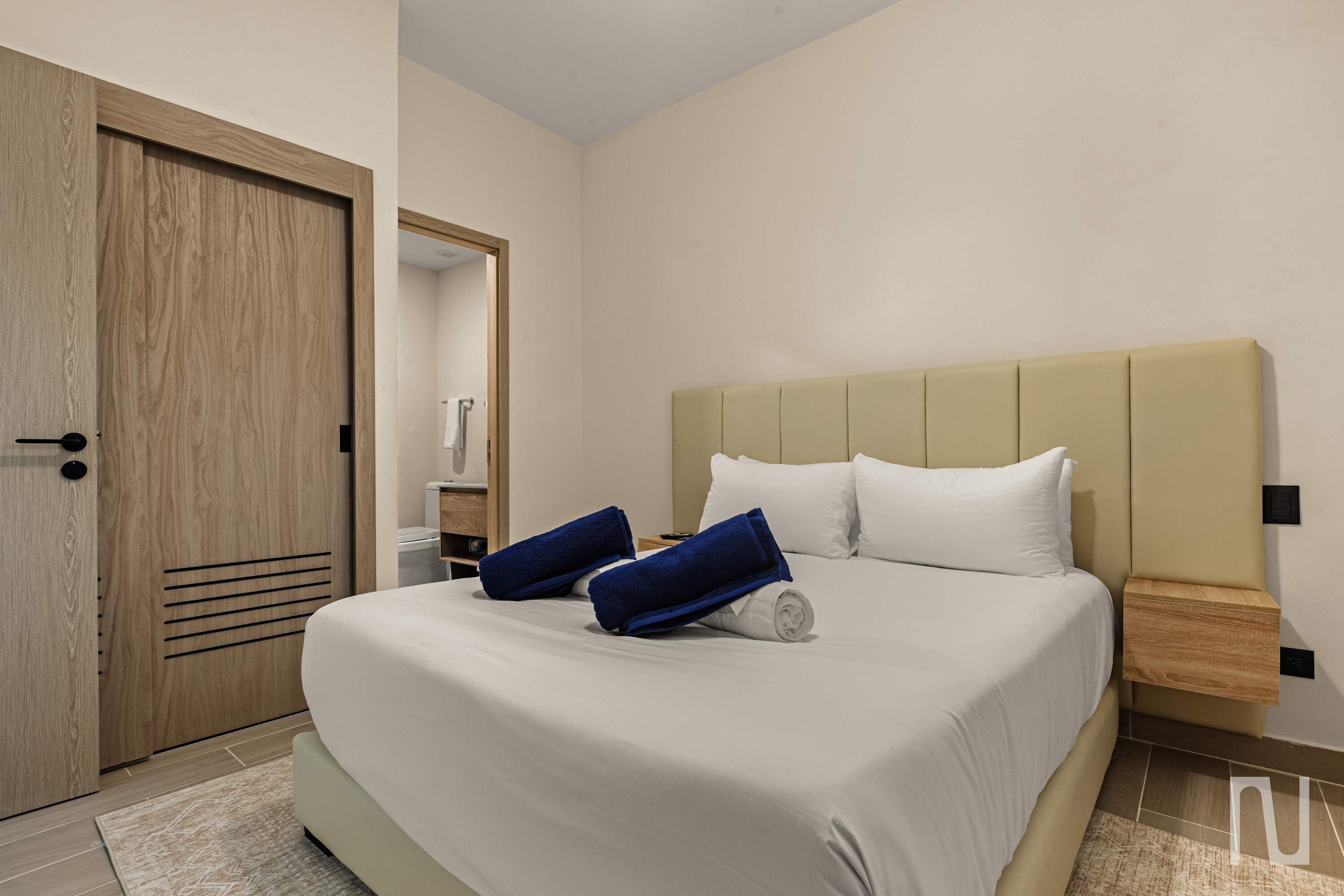 A hotel room with a bed, a wooden door, a nightstand, and a bathroom visible through an open door.