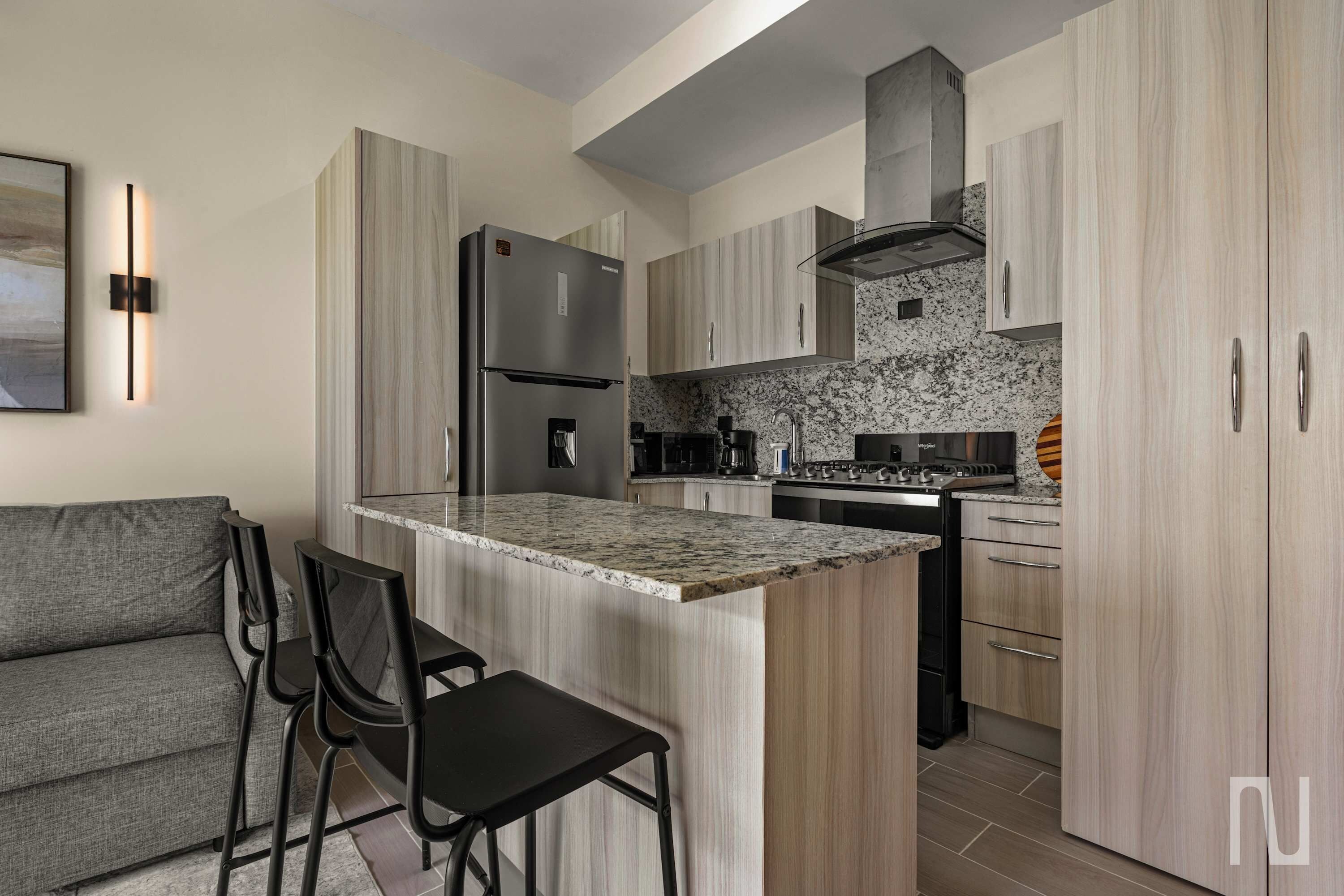 A modern kitchen with a central island, stainless steel appliances, and a grey sofa with black chairs.