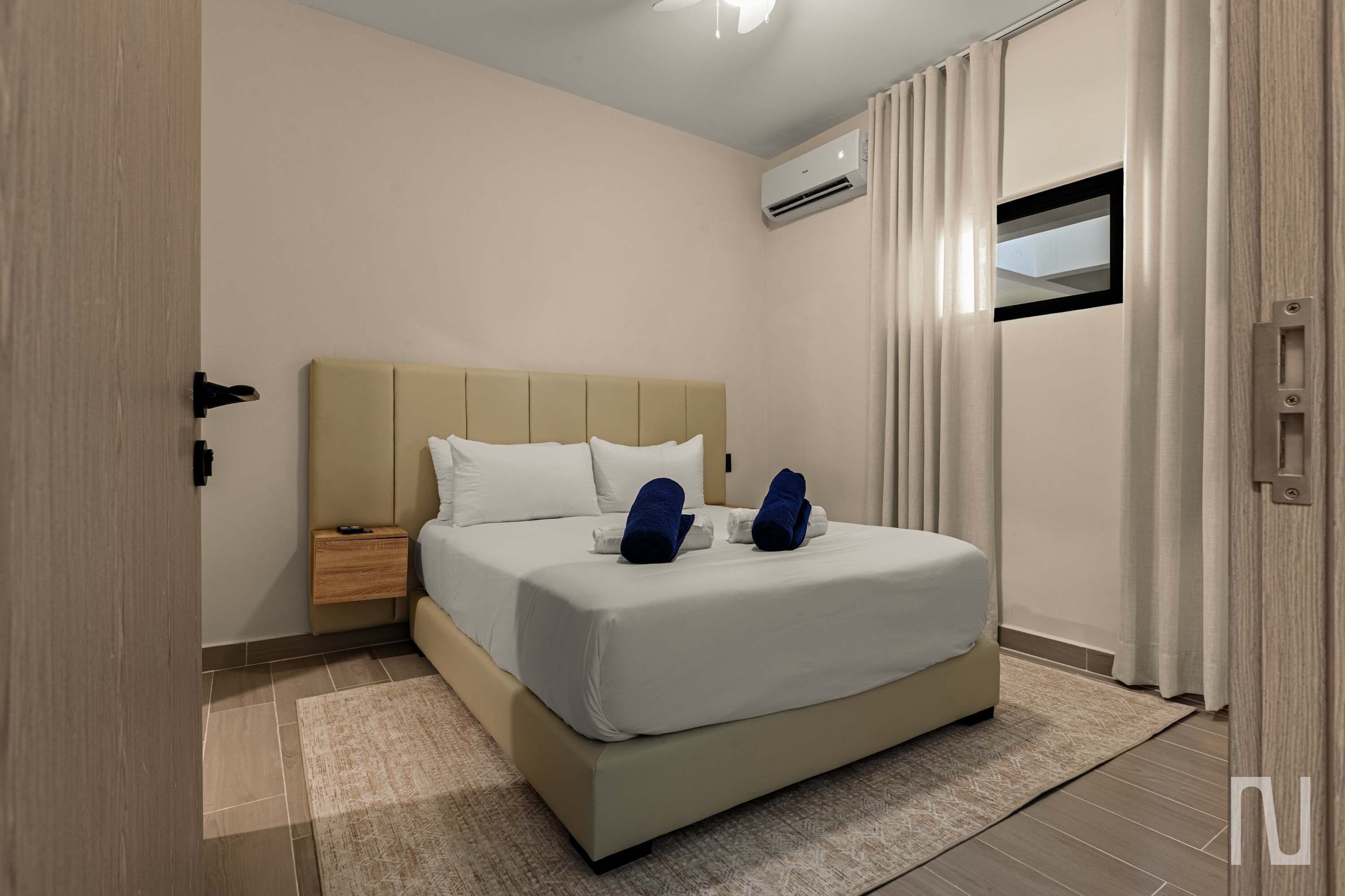 A hotel room with a bed, a nightstand, an air conditioning unit, and a window with curtains.