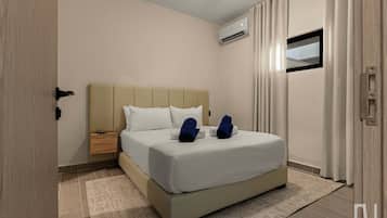 A hotel room with a bed, a nightstand, an air conditioning unit, and a window with curtains.