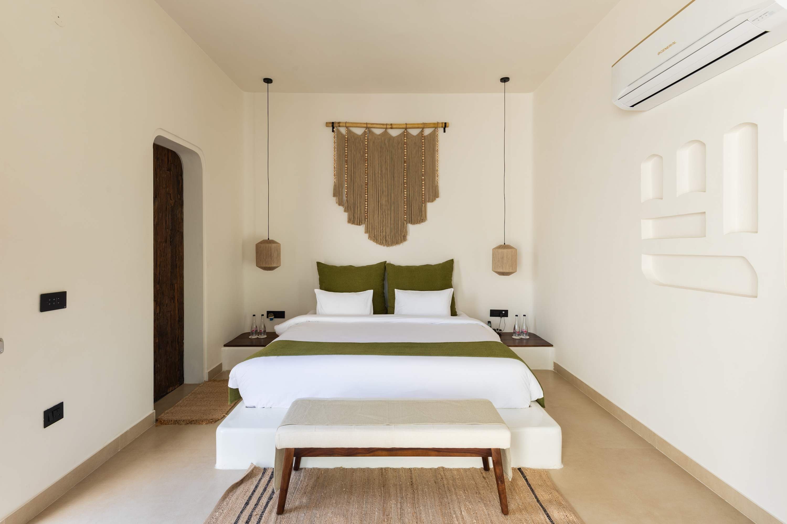 A bedroom with a bed, bedside tables, a bench, a wall-mounted air conditioner, and a door.
