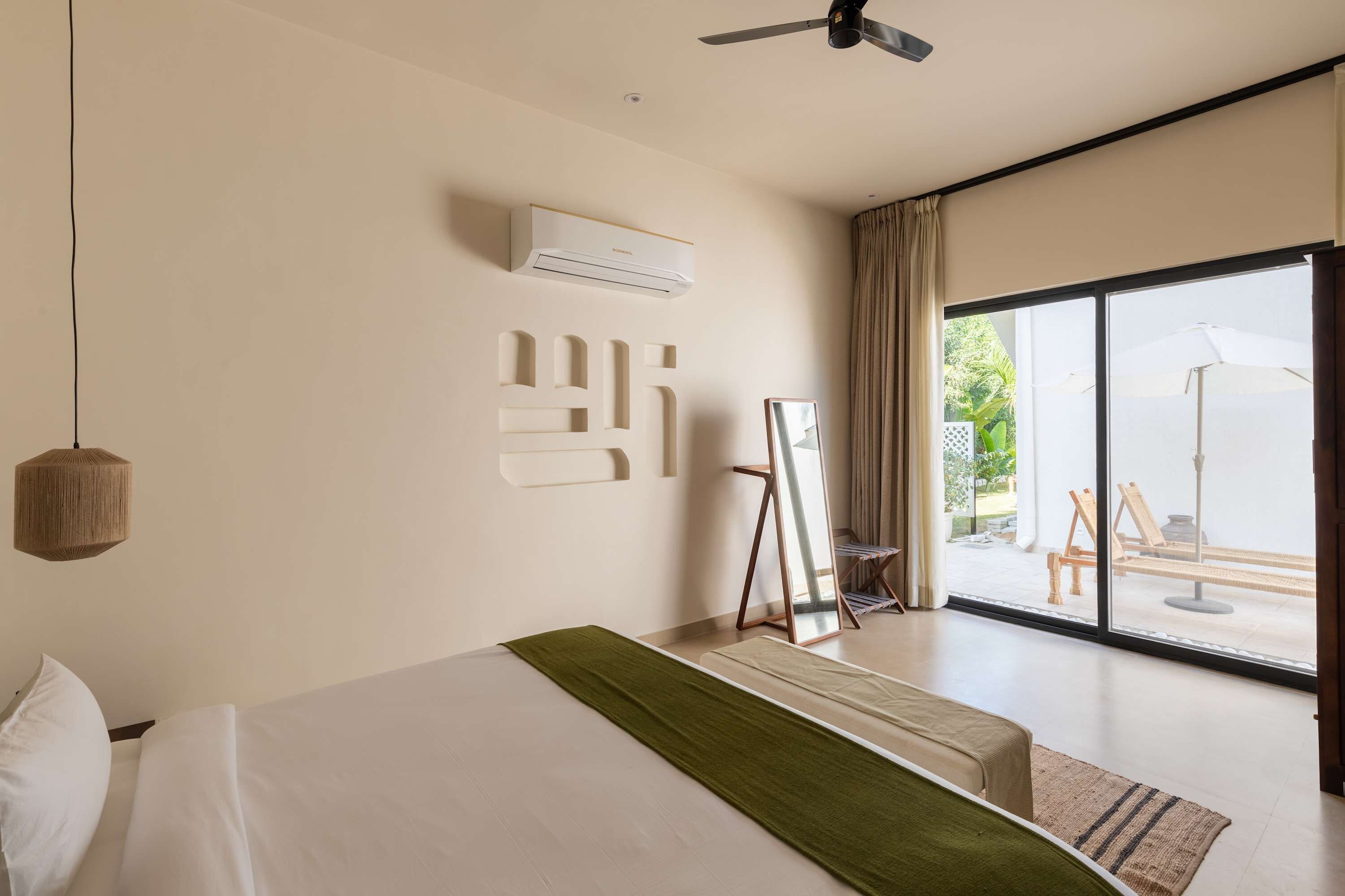 A hotel room with a bed, a ceiling fan, an air conditioning unit, a large window with a view of the outdoors, and a painting on the wall.