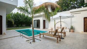 Un espace piscine doté d’un toit de chaume, de chaises longues et d’un parasol blanc.