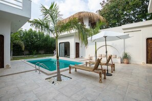 Un espace piscine doté d’un toit de chaume, de chaises longues et d’un parasol blanc.