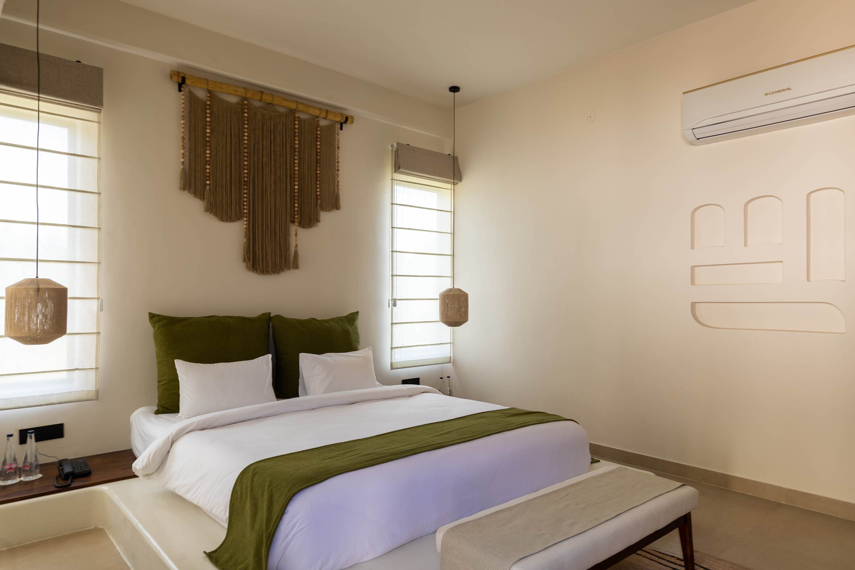 A hotel room with a bed, bedside tables, a bench, and a wall-mounted air conditioning unit.