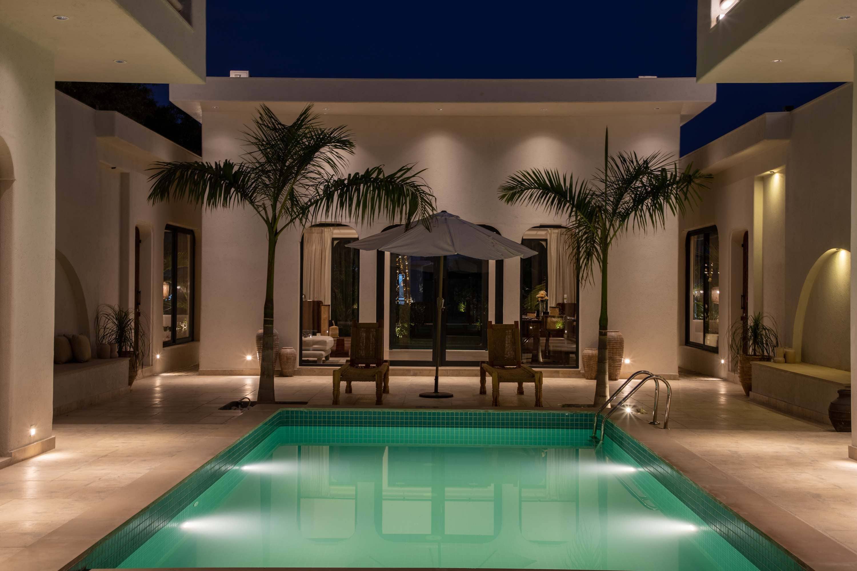Un área de piscina interior moderna con una piscina cristalina, rodeada de palmeras y una zona de estar con sillas y una mesa.
