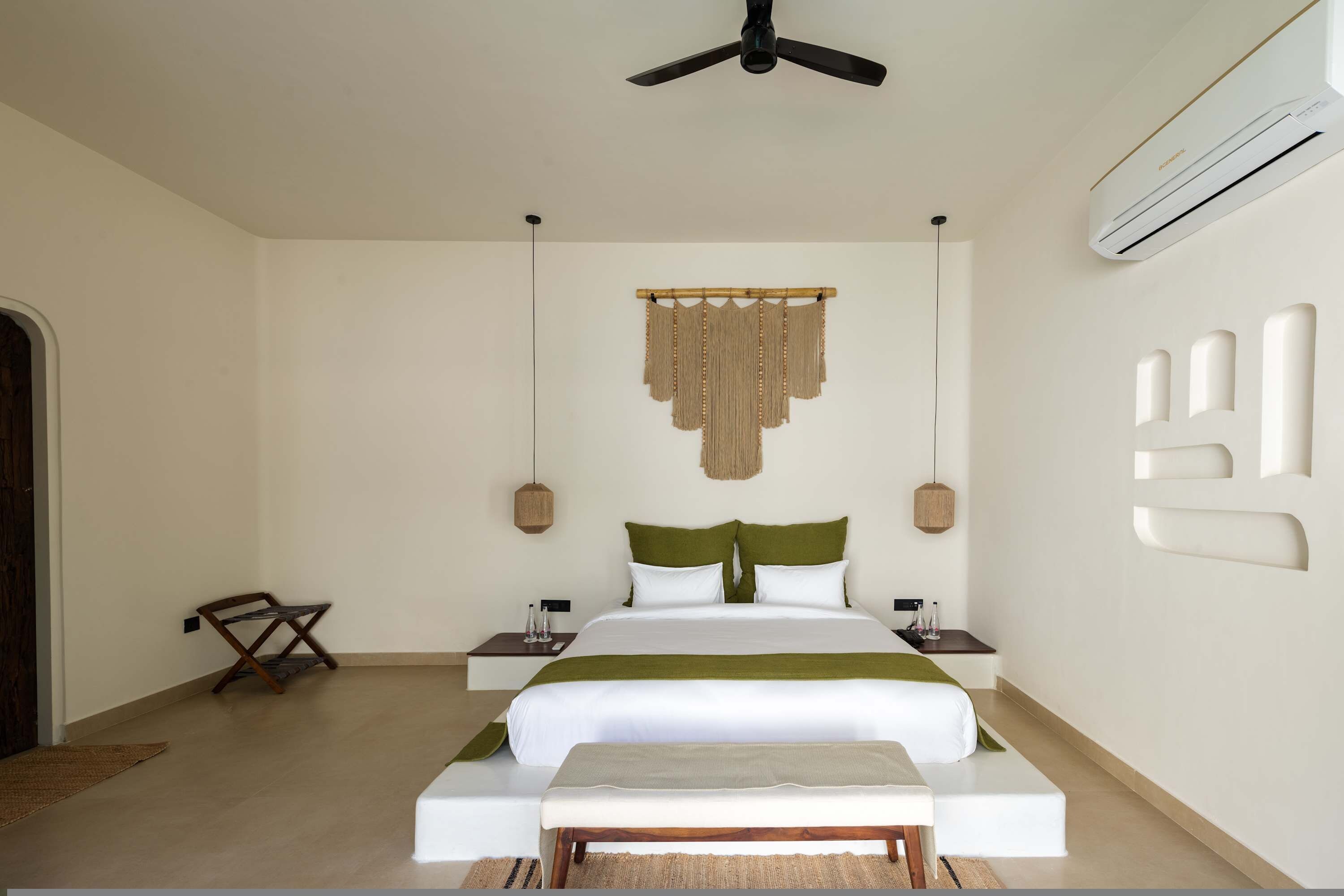 A hotel room with a bed, bedside tables, a hanging macrame wall decoration, a ceiling fan, and an air conditioning unit.