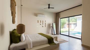 A modern bedroom with a bed, a ceiling fan, an air conditioning unit, a window with a view of a swimming pool, and a framed picture on a stand.