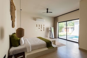 A modern bedroom with a bed, a ceiling fan, an air conditioning unit, a window with a view of a swimming pool, and a framed picture on a stand. - La Vida Retreat - Luxury 4 Bed Villa in Jaipur (Jaipur)
