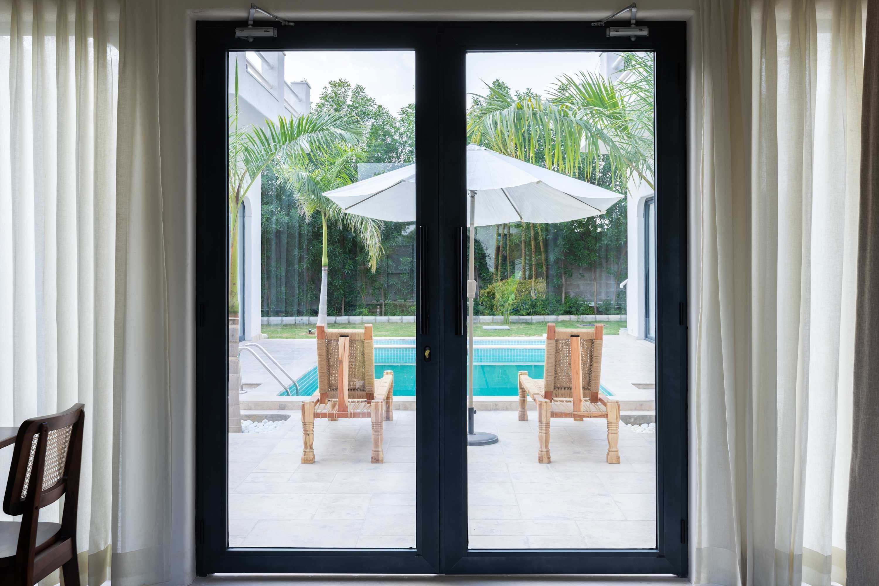 On aperçoit, à travers une porte vitrée, un espace piscine avec deux chaises en bois et un parasol blanc.