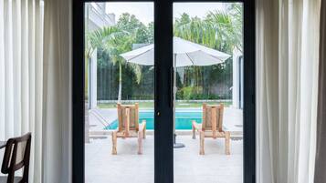 On aperçoit, à travers une porte vitrée, un espace piscine avec deux chaises en bois et un parasol blanc.
