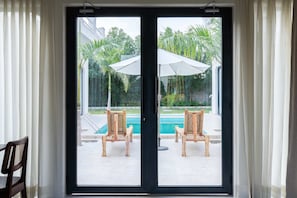 On aperçoit, à travers une porte vitrée, un espace piscine avec deux chaises en bois et un parasol blanc.