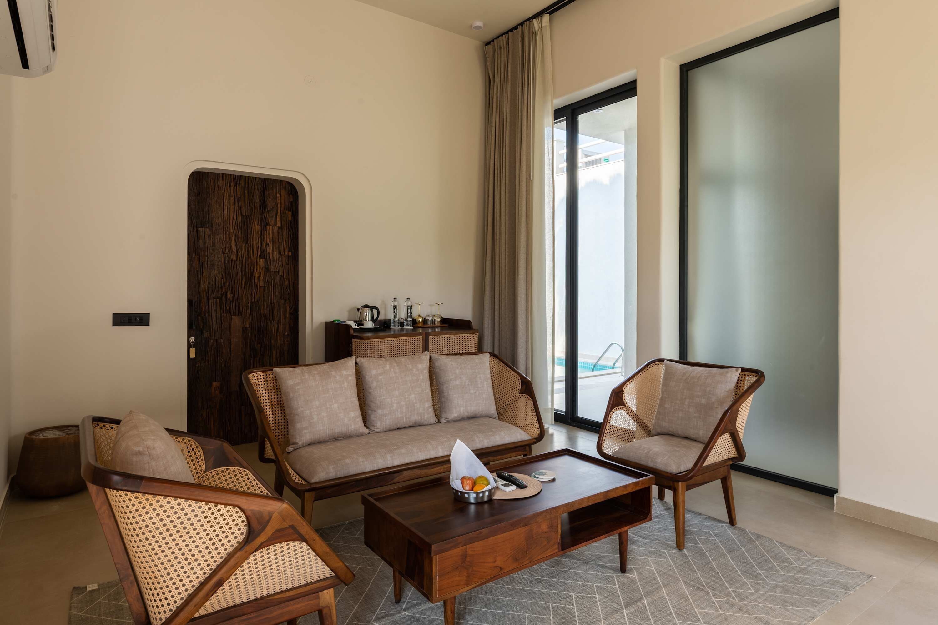 A modern living room with a sofa, two armchairs, a coffee table, and a dining table. There is a large window with a view of a pool.