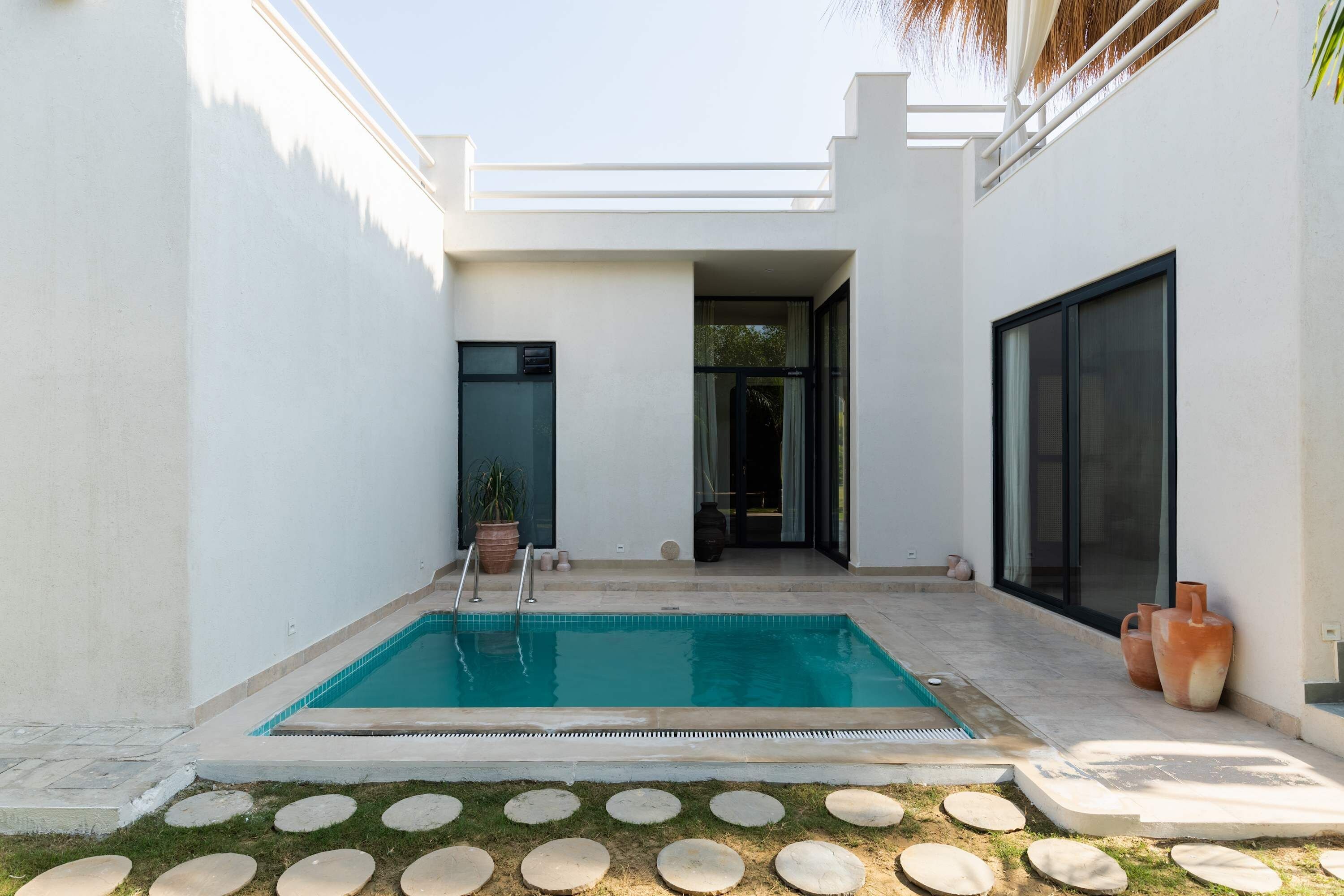 Un área de piscina moderna al aire libre con una piscina azul cristalina, rodeada por un muro blanco y un pequeño jardín con piedras para caminar.