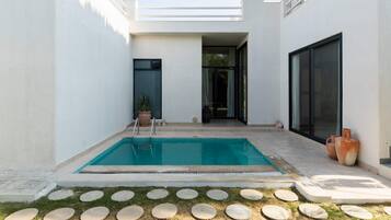 Un área de piscina moderna al aire libre con una piscina azul cristalina, rodeada por un muro blanco y un pequeño jardín con piedras para caminar.