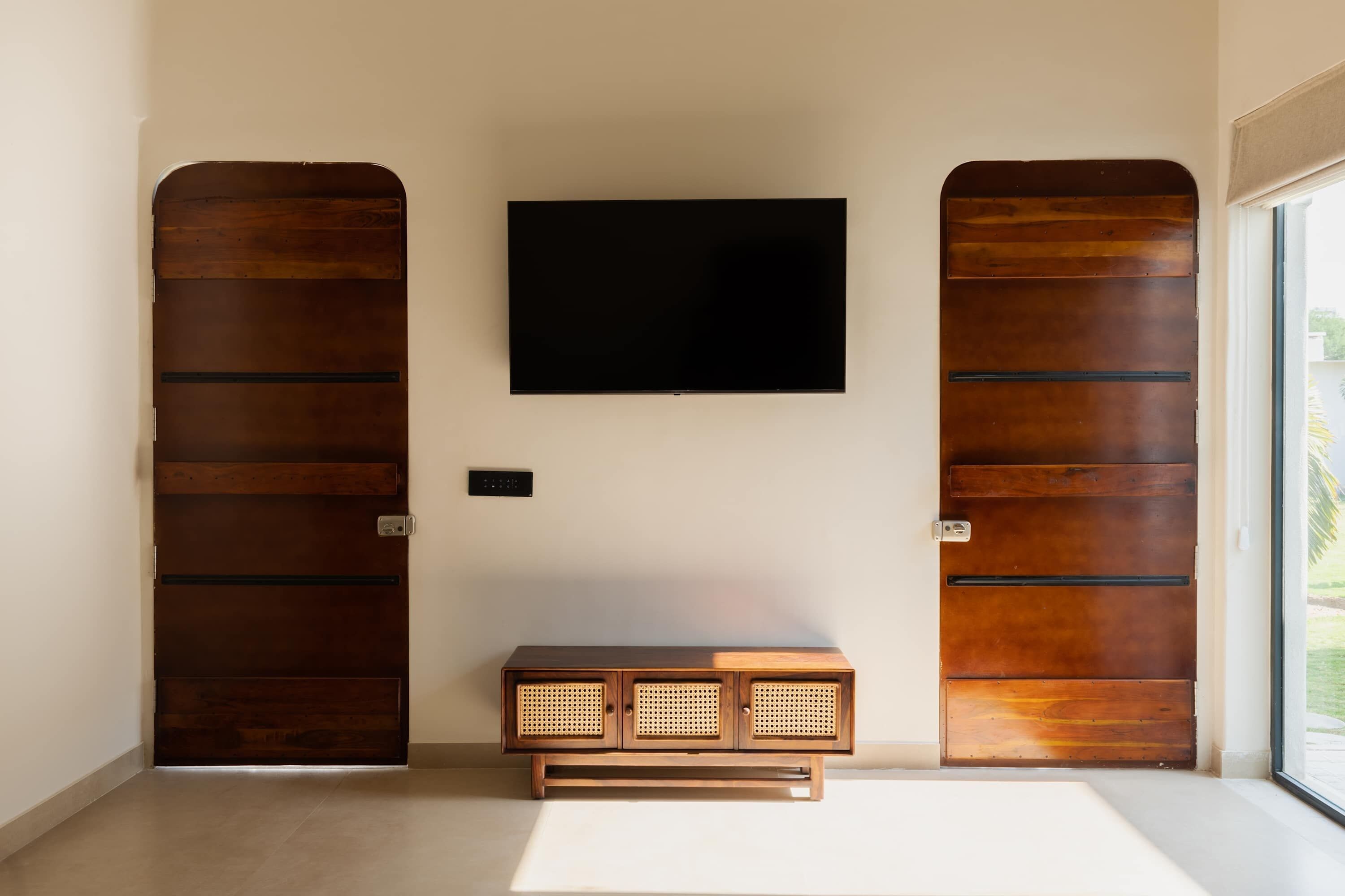 Una habitación con una televisión de pantalla plana grande montada en la pared, un mueble de madera con frente de ratán y dos puertas de madera con pomos de metal.