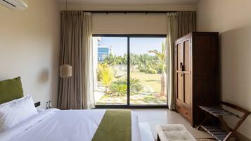Habitación de hotel con cama, armario de madera, banca y vista a un jardín y un edificio a través de la ventana.