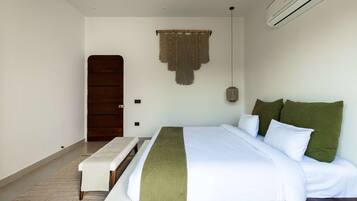 A bedroom with a bed, bedside table, bench, wall-mounted air conditioning unit, and a door.