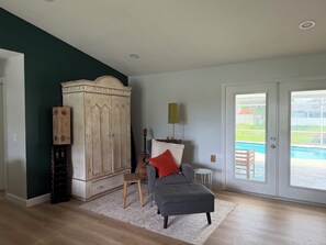 A room with a large wooden wardrobe, a grey sofa with a red pillow, a small wooden stool, and a guitar. There is a white lamp and a white rug on the floor.