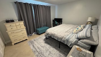 Quarto com cama, penteadeira, banquinho, abajur e pequena mesa com copo e vela.