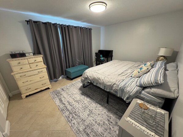 A bedroom with a bed, dresser, bench, lamp, and a small table with a glass and a candle.
