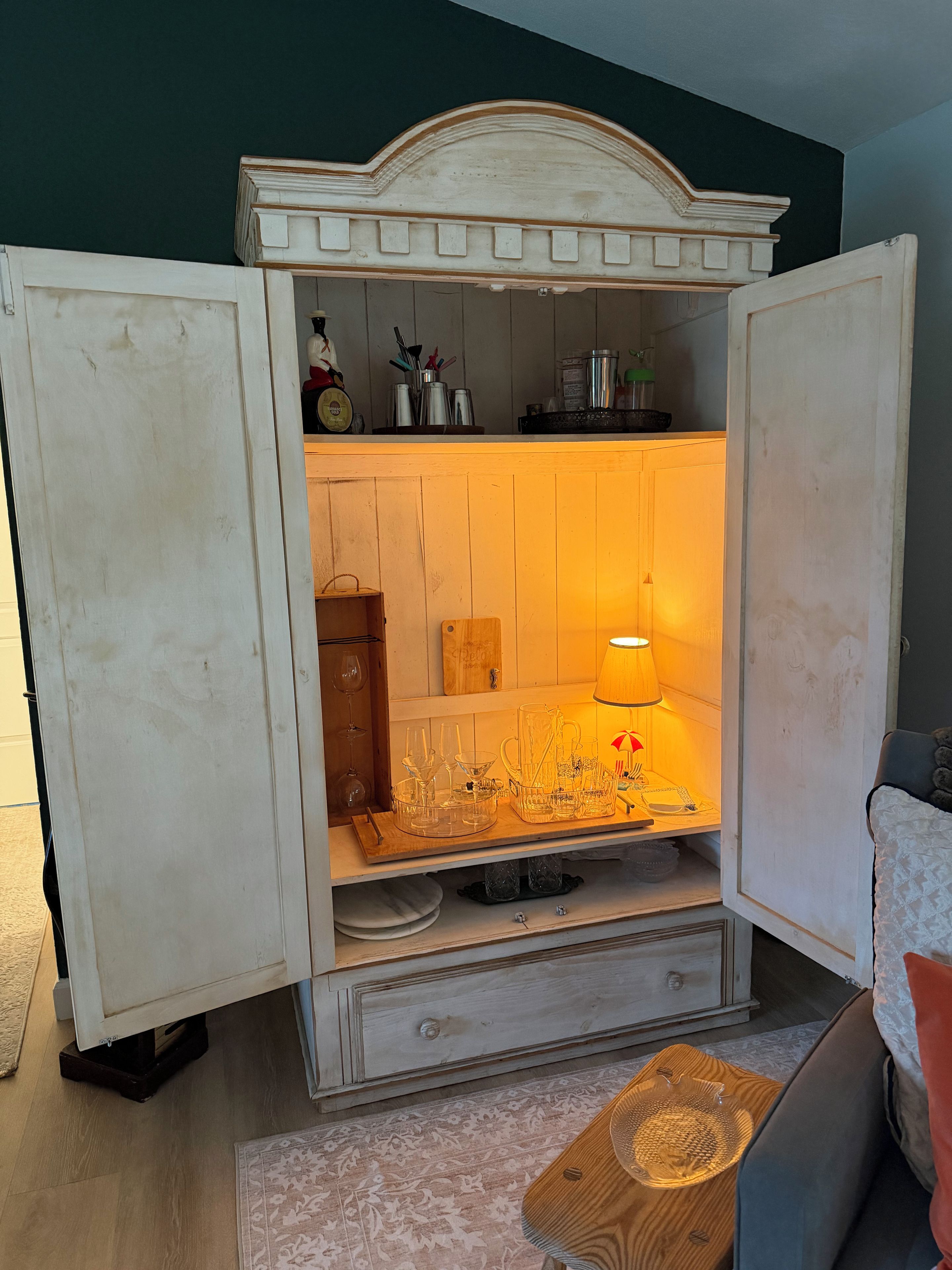 A room with a vintage-style cabinet with open doors, revealing a shelf with various items and a lit lamp.