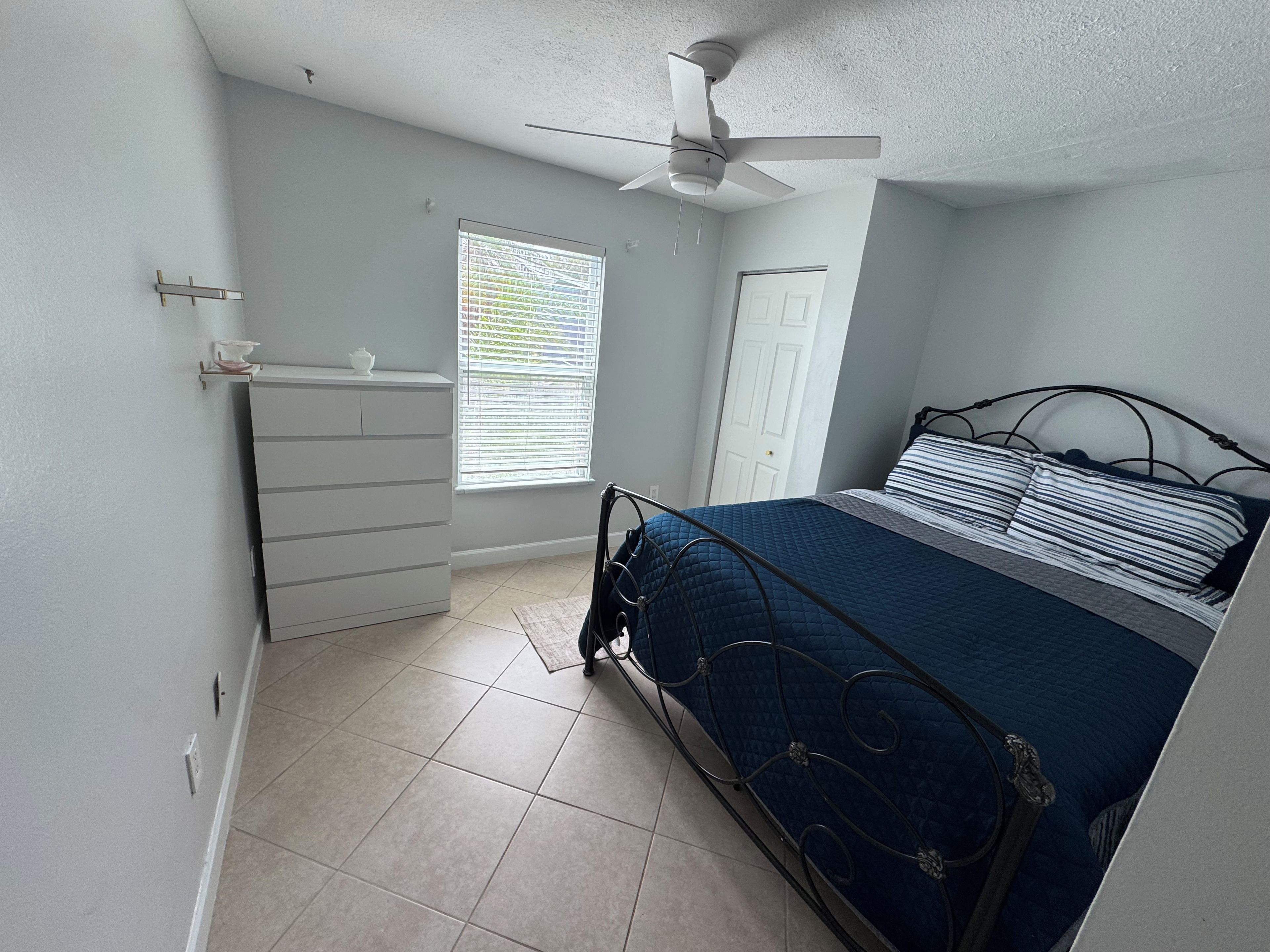 A bedroom with a bed, a dresser, a ceiling fan, a window with blinds, and a door.